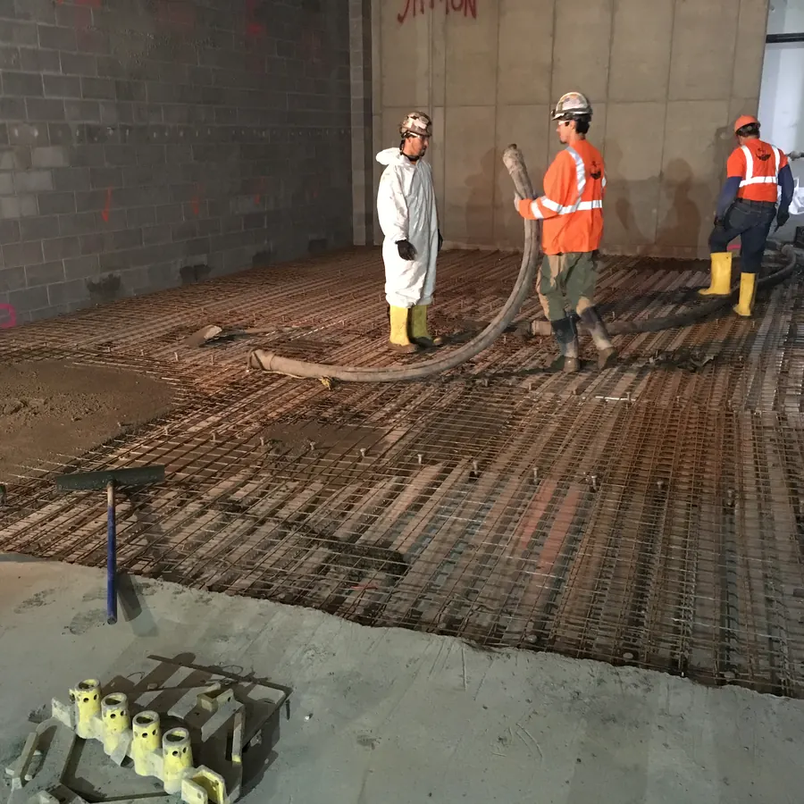 Tiga pekerja konstruksi dengan perlengkapan APD keselamatan dan sepatu bot sedang memegang selang pompa beton untuk mengecor plat lantai dak beton bertulang di atas anyaman besi tulangan