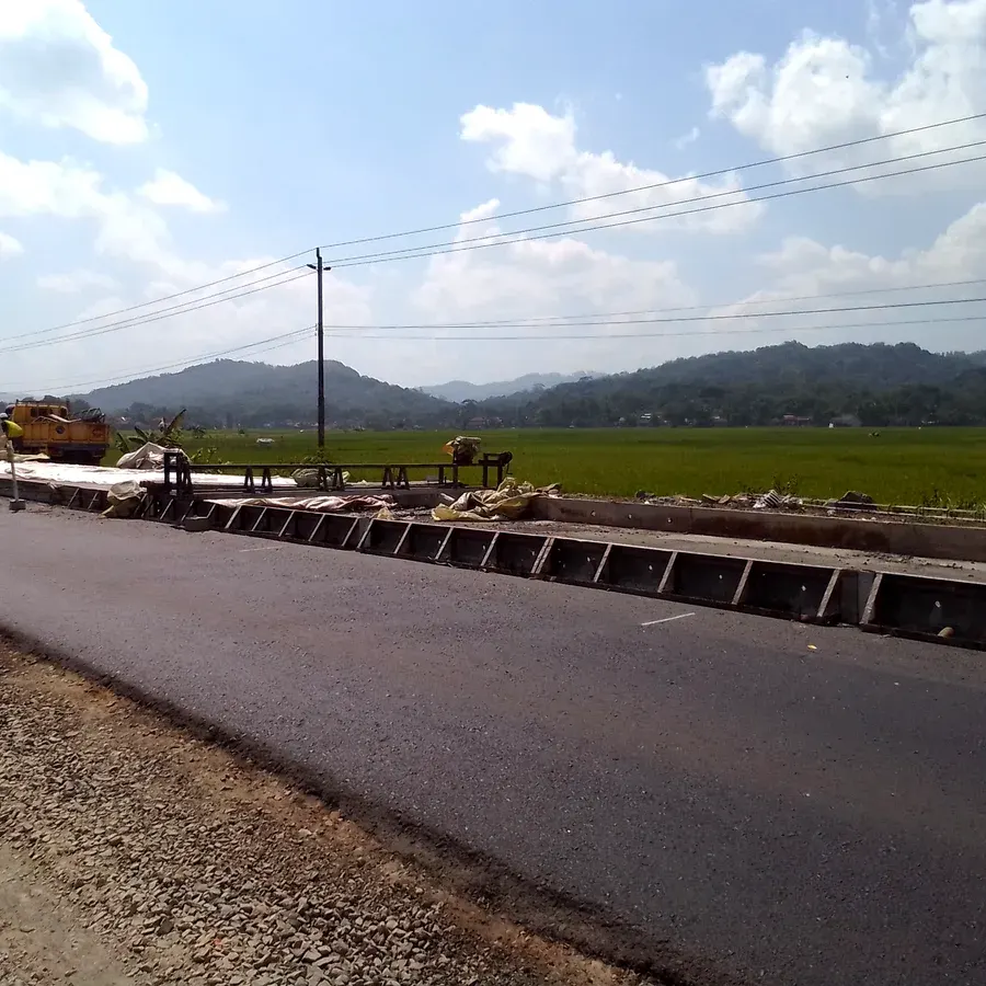 Pengerjaan proyek jalan dengan cetakan bekisting cor beton baja dan hamparan agregat lapis pondasi di tepi hamparan sawah hijau berlatar perbukitan