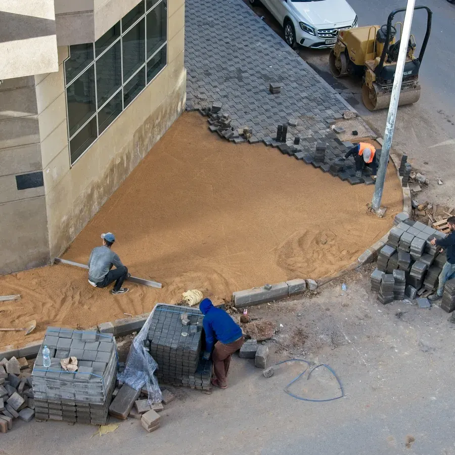 Pekerja konstruksi sedang meratakan hamparan pasir alas bedding sand menggunakan jidar logam dan memasang paving block conblock persegi panjang di trotoar tepi jalan