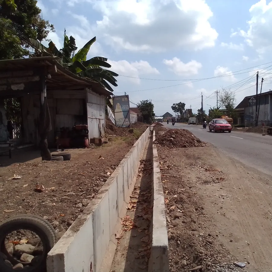 Pemandangan proyek pembuatan saluran drainase beton pracetak di bahu jalan aspal dengan gundukan tanah galian dan timbunan agregat di tepi jalan lingkungan