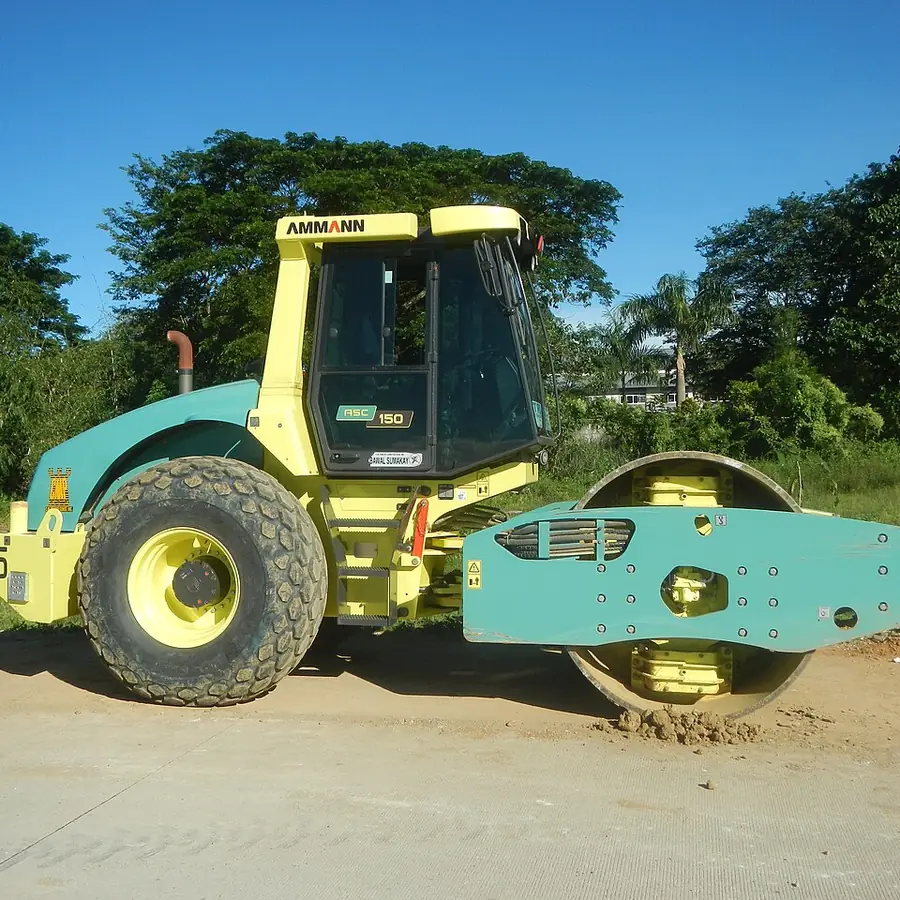 Alat berat vibro road roller compactor Ammann beroda drum baja dan ban alur tebal sedang memadatkan lapisan tanah timbunan di tepi jalan dekat pepohonan tropis